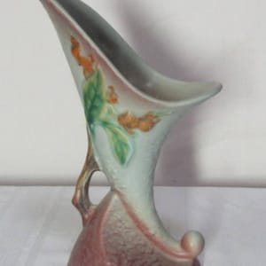 Puede incluir: Un jarrón de cerámica artística vintage con un diseño asimétrico único. El jarrón presenta un cuerpo azul claro con una abertura acampanada y una base curva. Está adornado con detalles florales naranjas y verdes y un asa. La base es de color rosa apagado.