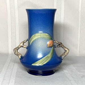 Puede incluir: Un jarrón de cerámica azul con cuerpo bulboso y parte superior acampanada. El jarrón presenta dos asas en forma de rama y un diseño floral pintado en verde y naranja. El jarrón mide aproximadamente 20 cm de alto.