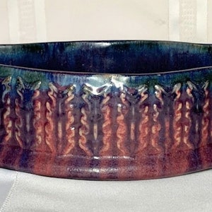 May include: A handmade ceramic planter with a deep purple, blue, and green glaze. The planter has a wavy rim and a decorative pattern of stylized leaves around the bottom.