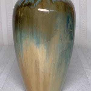 May include: A ceramic vase with a glossy finish. The vase has a dark green rim and a body that transitions from tan to blue-green. The colors blend together, creating a unique, artistic look. The vase is a decorative item.