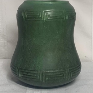 Puede incluir: Un jarrón verde con una superficie texturizada y un patrón geométrico alrededor de la parte superior e inferior. El jarrón tiene una forma redondeada, más ancha en la base y en la parte superior, y más estrecha en el medio.