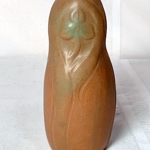 May include: A brown ceramic vase with a stylized floral design. The vase has a narrow neck and a rounded body. The floral design is in a lighter shade of brown with a touch of green. The vase is approximately 8 inches tall.