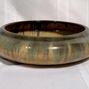 May include: A ceramic bowl with a wide, rounded rim. The bowl's exterior features a blend of earthy tones, including shades of green, brown, and tan. The interior has a dark brown glaze. The bowl is sitting on a white surface.