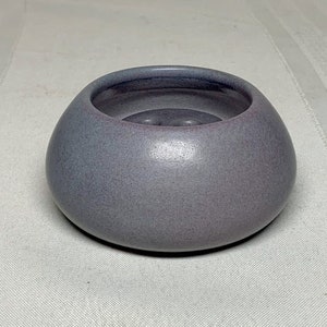 Pode incluir: Uma pequena tigela redonda de cerâmica com um acabamento fosco salpicado. A tigela é de cor roxa suave com um interior roxo mais escuro e uma borda ligeiramente avermelhada. A tigela tem uma forma lisa e arredondada.