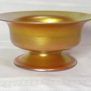 May include: A gold iridescent glass bowl with a pedestal base. The bowl has a smooth, rounded shape and a slightly flared rim.