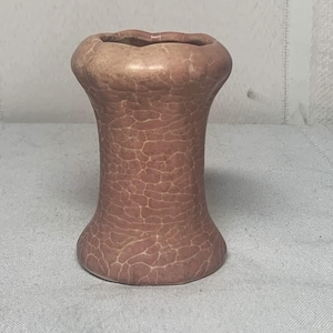 Puede incluir: Un pequeño jarrón decorativo con una superficie texturizada y agrietada en tonos rosa y beige. El jarrón tiene una base acampanada, un cuerpo cilíndrico y un borde suavemente festoneado. Este jarrón de cerámica es adecuado para exhibir flores pequeñas.