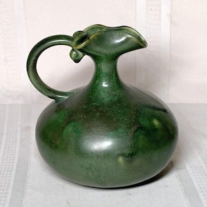 May include: A green ceramic pitcher with a wide, rounded body and a flared spout. The pitcher has a unique, leaf-shaped handle.