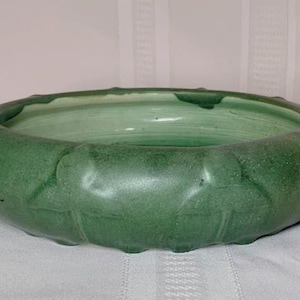 Puede incluir: Un cuenco de cerámica verde con un exterior texturizado y redondeado. El cuenco tiene un diseño ancho y poco profundo con un borde ligeramente irregular. El interior tiene un tono verde más claro. Este cuenco decorativo es adecuado para diversos usos.