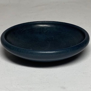 May include: A circular, shallow bowl with a smooth, matte finish in a deep, dark navy blue color. The bowl features a thick, rounded rim and a concave center, presenting a simple, functional design with a consistent, solid dark blue tone throughout.