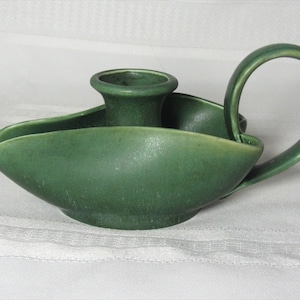 Puede incluir: Un portavelas de cerámica verde con un solo portavelas y un asa. El portavelas tiene forma de barco.