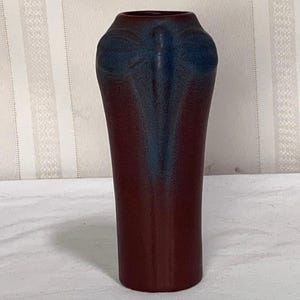 May include: A cylindrical ceramic vase with a matte finish, featuring a deep burgundy base color that transitions into a dark blue gradient at the top. The upper section is embossed with a stylized dragonfly motif with wings extending outward.