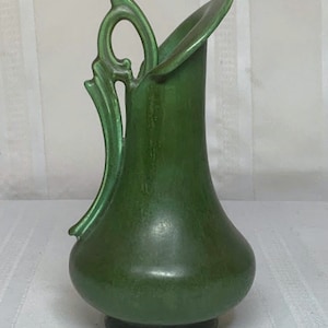 Puede incluir: Una jarra de cerámica verde con un asa adornada. La jarra tiene un acabado liso y brillante y un cuerpo redondeado.