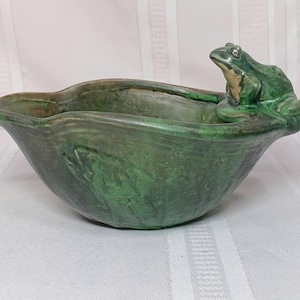 May include: Green ceramic bowl with a frog figurine perched on the rim. The bowl has a textured surface and a leaf design. The frog is green and tan, adding a whimsical touch to the decorative piece.
