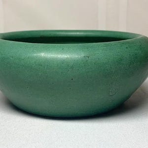 May include: A round, green ceramic bowl with a wide opening. The bowl has a smooth, matte finish and a slightly textured surface. The color is a muted, earthy green. This bowl is suitable for decorative or functional use.