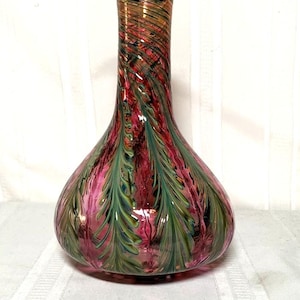 May include: A decorative glass vase with a bulbous base and a slender neck. The vase features a vibrant color palette of pink, green, and gold, with a swirling, leaf-like pattern. The rim is a deep blue.