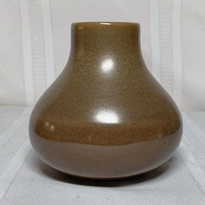 Könnte beinhalten: Eine braune Keramikvase mit rundem Körper und schmalem Hals. Die Vase hat eine glänzende Oberfläche mit einem dunkleren braunen Sockel und einem helleren braunen Oberteil. Die Vase steht auf einer weißen Oberfläche.