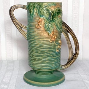 May include: A green ceramic vase with a pedestal base and two curved handles. The vase features a textured surface with embossed grape leaves and bunches of grapes. The handles are a contrasting brown color.