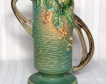 May include: A green ceramic vase with a pedestal base and two curved handles. The vase features a textured surface with embossed grape leaves and bunches of grapes. The handles are a contrasting brown color.