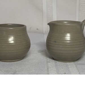 May include: Two matching stoneware pieces in a neutral beige color. One is a small, round, open container. The other is a small pitcher with a handle and a spout. Both have horizontal ridged detailing.