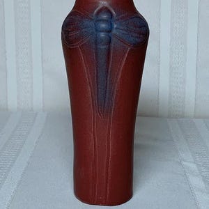 Puede incluir: Un jarrón alto y rojo con un diseño de libélula en azul y morado. El jarrón tiene una forma delgada y alargada y una superficie texturizada. La libélula está centrada en la parte delantera del jarrón, con las alas extendidas. El jarrón es probablemente de cerámica o alfarería.