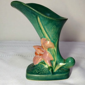 May include: A green glazed ceramic vase with a unique shape and a floral design. The vase features a pink and orange flower with green leaves.