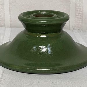 Puede incluir: Un portavelas de cerámica verde con una base ancha y un centro elevado para la vela. El portavelas tiene un acabado brillante y un diseño sencillo. La forma general recuerda a un cuenco pequeño.