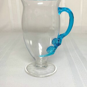 May include: A clear glass mug with a blue rim and handle. The handle is shaped like a seashell.