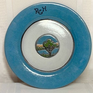 Könnte beinhalten: Ein blauer Keramikteller mit einer weißen Mitte, auf der eine gemalte Szene mit einem Baum und einem See dargestellt ist. Der Teller trägt die Initialen "RGH" in Schwarz am Rand.