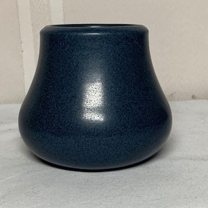 May include: A dark blue ceramic vase with a smooth, rounded silhouette and a wide, curved base that tapers toward a narrow, circular opening at the top. The surface features a subtle, speckled texture with a matte finish and a light reflection.