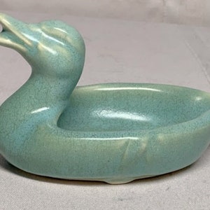 Pode incluir: Um prato de cerâmica azul claro em forma de pato. A cabeça do pato está levantada e o prato tem uma forma oval pouco profunda.