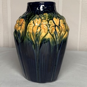 May include: A ceramic vase featuring a deep navy blue glaze with a glossy finish. The upper portion of the vase is decorated with a raised, molded relief pattern of yellow iris flowers and green foliage that transitions into the dark blue base.