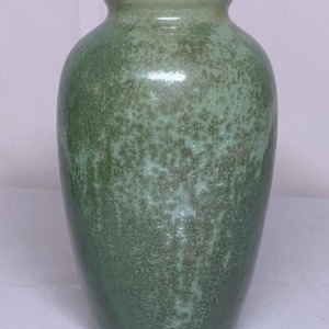 May include: A green ceramic vase with a speckled texture. The vase has a rounded body and a slightly flared rim. The color is a muted green with darker green speckles throughout. The vase is approximately 8 inches tall.