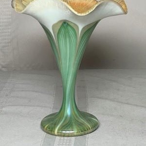 May include: A decorative glass vase with a ruffled rim. The vase features a white interior, a green and gold patterned body, and a ruffled rim with orange and cream accents. The vase has a flared base and a slender, elongated neck.