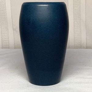 May include: A cylindrical vase with a smooth, matte finish in a deep, solid navy blue color. The vessel features a slightly tapered base and a wide, open top, presenting a clean, uniform appearance with no additional patterns or decorative elements.