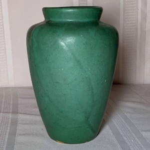 Può includere: Un vaso in ceramica verde con corpo arrotondato e un labbro leggermente svasato. Il vaso ha una finitura liscia e opaca.
