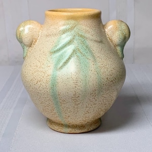 Pode incluir: Um vaso de cerâmica vintage com duas alças, com um esmalte verde claro e bege com um design de folhas. O vaso tem uma textura salpicada e um corpo arredondado.