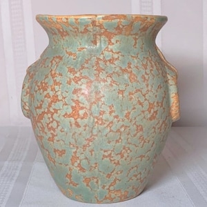 Puede incluir: Un jarrón de cerámica verde y marrón con un esmalte moteado y cuatro asas. El jarrón tiene un cuerpo redondeado y una boca ancha.