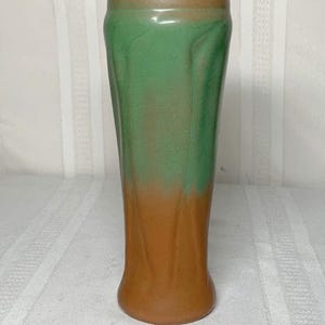 May include: A tall, ceramic vase with a two-tone color scheme. The top half is a muted green, transitioning to a warm brown at the base. The vase has a slightly curved, fluted design, adding visual interest to its form. The vase is approximately 10 inches tall.