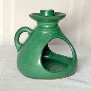 Può includere: Un portacandele in ceramica verde con un design unico. Ha una grande apertura al centro e un manico sul lato. Il portacandele è smaltato con una finitura liscia e lucida.