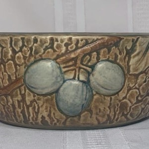 May include: A ceramic bowl with a textured, bark-like exterior in shades of brown and tan. The bowl features a branch with three blue-green fruit. The design is embossed, adding depth and detail to the decorative piece.