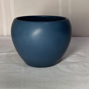 May include: A deep navy blue ceramic bowl with a smooth, matte finish and a rounded, bulbous silhouette. The vessel features a wide, open circular top and a curved body that tapers slightly toward the base, presenting a solid, uniform color.