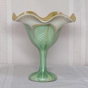 May include: A decorative glass pedestal bowl with a ruffled rim. The bowl features a green and white swirled pattern on the body and a gold-toned rim. The base and stem are green. The bowl is likely used for serving or display.