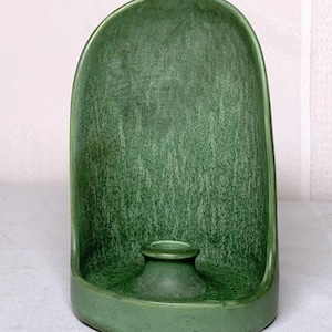 Puede incluir: Un portavelas de cerámica verde con una parte trasera curvada y una pequeña copa circular para velas. El soporte tiene una superficie texturizada y una base redondeada. El diseño es simple y elegante, adecuado para la decoración del hogar.