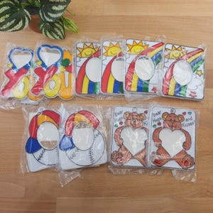 May include: A collection of rectangular plastic photo frames in individual clear packaging. Designs include colorful rainbows with suns, baseball caps, and brown teddy bears holding heart-shaped frames with the text Bear Hugs and Kisses.