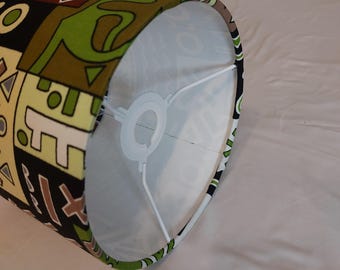 May include: A cylindrical lampshade with a colorful geometric pattern in green, brown, black, and cream. The interior is white with a metal frame. The lampshade is resting on a white surface.