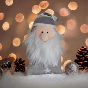 May include: A plush Christmas gnome with a white faux fur beard, a pink nose, and a gray and white striped hat. The gnome is wearing a gray outfit with a white star. The background features bokeh lights and decorative elements.