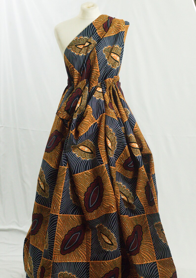 May include: A one-shoulder dress made from a colorful African print fabric. The fabric features a geometric pattern of orange, brown, and blue with a white outline. The dress is gathered at the waist and has a flowing skirt.