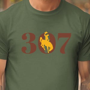May include: Olive green t-shirt featuring the number "307" in a dark red font, with a yellow silhouette of a cowboy riding a bucking bronco incorporated into the zero. The shirt is made of a soft material.