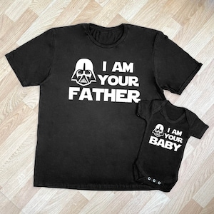 Peut inclure: Deux t-shirts noirs avec un motif blanc de Dark Vador et le texte "Je suis ton père" sur le plus grand t-shirt et "Je suis ton bébé" sur le plus petit.