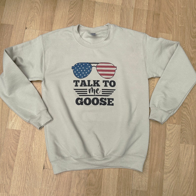 May include: A light beige sweatshirt with a graphic design of sunglasses with an American flag pattern. The text "TALK TO me GOOSE" is printed below the sunglasses.